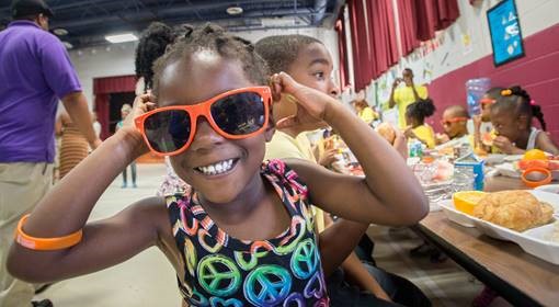 A smiling girl with orange glasses at lunch provided through assistance from the U.S. Department of Agriculture's (USDA) Food Nutrition Service (FNS). (Photo courtesy of the U.S. Department of Agriculture)