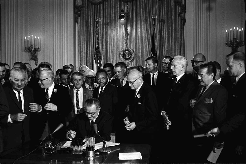 Lyndon B. Johnson signs the 1964 Civil Rights Act as Martin Luther King, Jr., others look on.